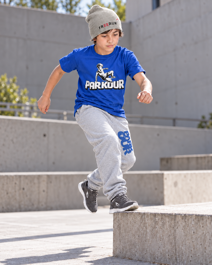 Blå Traceur T-shirt og grå/blå bukser med grå FREERUN-hue – under parkourtræning. Sko købes separat.