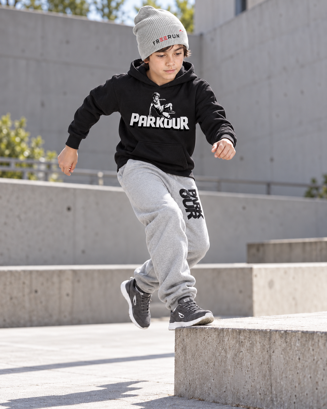 Sort Traceur-hoodie, grå/sorte bukser og grå FREERUN-hue – under parkourtræning.