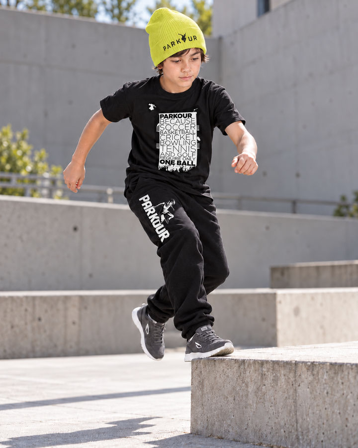 Sort Parkour Takes BALLS T-shirt, Traceur-bukser og gul Parkour-hue – under parkourtræning.